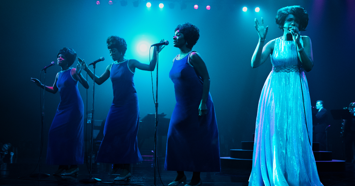 vestita di bianco e su palco illuminato di blu, aretha canta accanto a tre coriste - nerdface