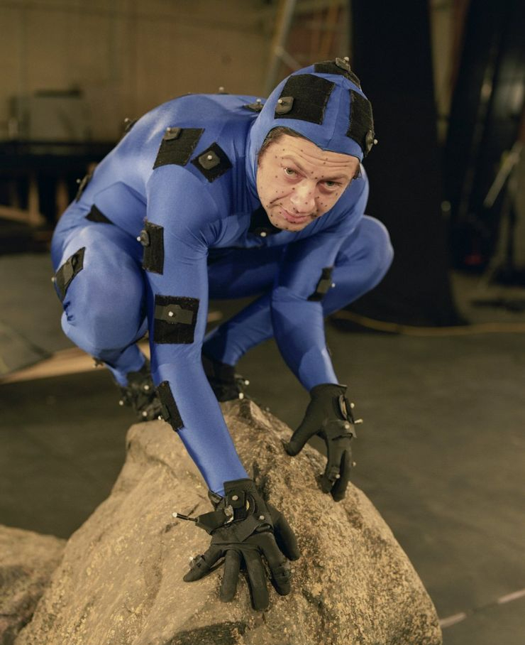 andy serkis in tuta blu è appollaiato su una roccia e interpreta gollum - nerdface