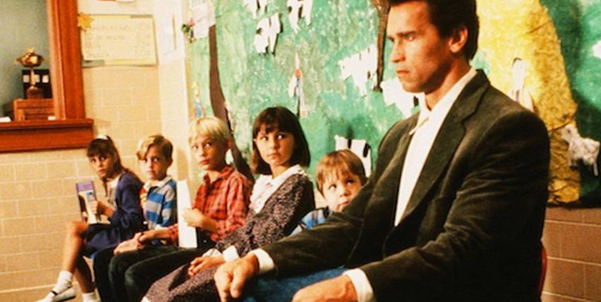 schwarzenegger è seduto serissimo e accanto diversi bambini lo guardano incuriositi in kindergarten cop - nerdface