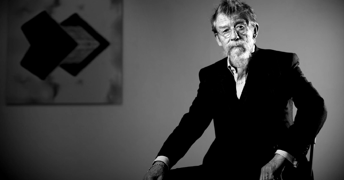 john hurt in una posa seria, seduta su una sedia e con un quadro di sfondo - nerdface