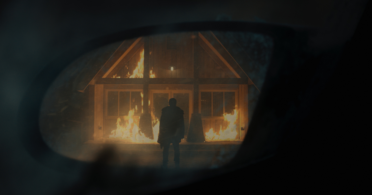 dal riflesso di uno specchietto retrovisore, si vede una figura stagliarsi di fronte un edificio in fiamme - nerdface