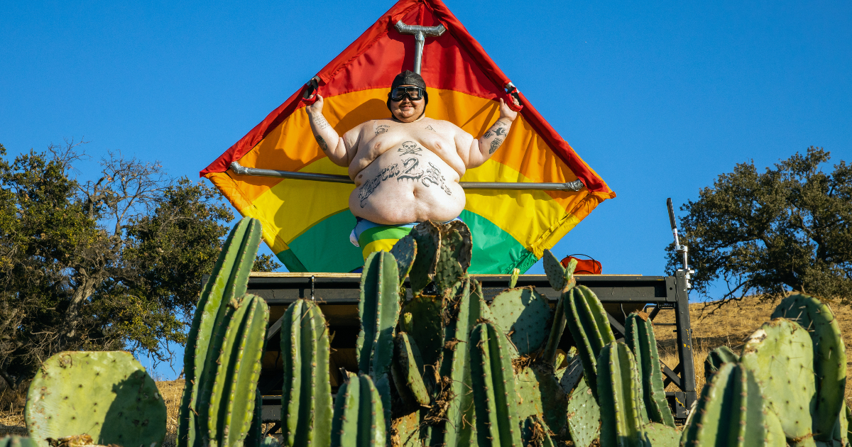 un uomo obeso a torso nudo legato a un enorme aquilone arcobaleno sta per planare sopra un campo di cactus - nerdface